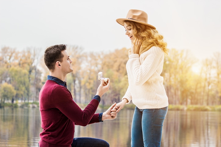 young man on one knee and giving engagement ring while making marriage proposal to his girlfriend outdoors.