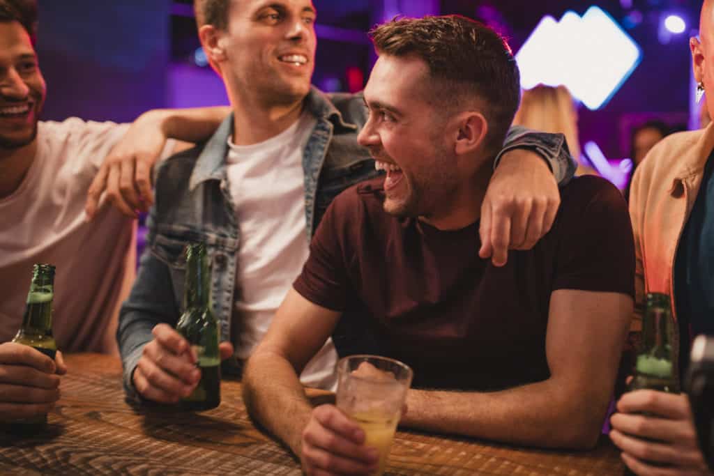 groomsmen at the bar with a drink each other, they are smiling and laughing at each other.